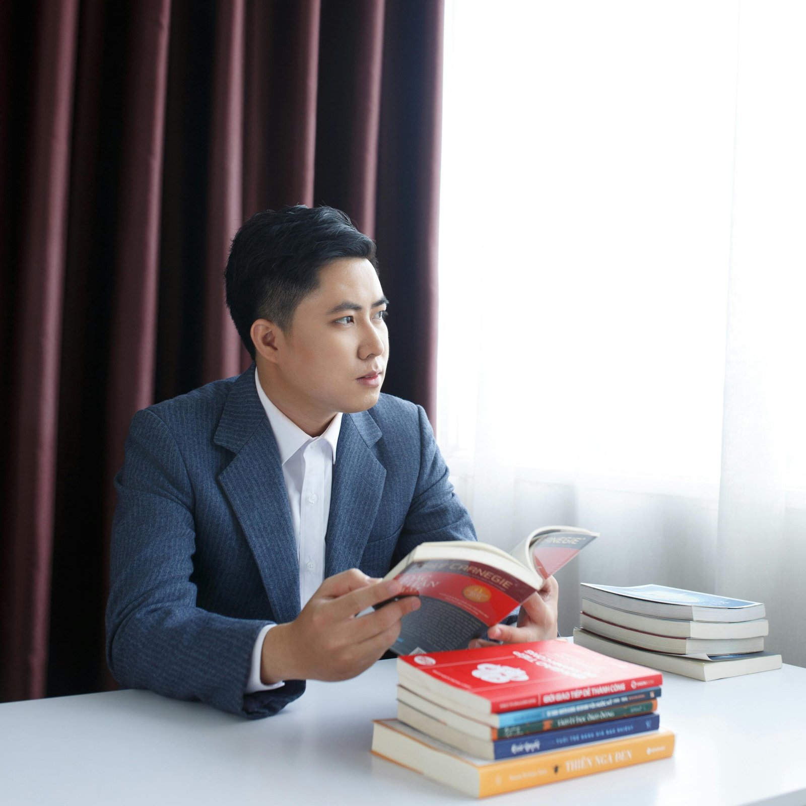 Young man in a suit reading a book at a table with a stack of books, in an indoor setting.