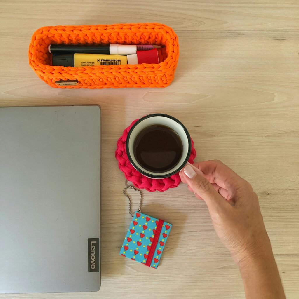 Woman's hand holding coffee on a vibrant desk with crochet container and Lenovo laptop.